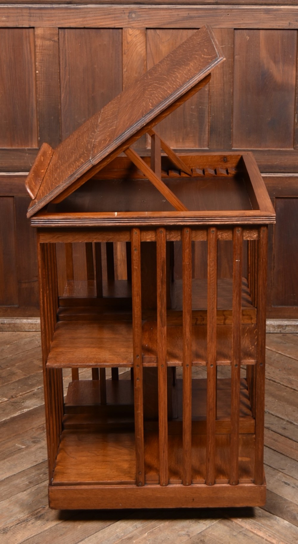 A two-tier freestanding timber revolving bookcase with slatted sides and an angled reading lectern top holding an open volume. View V