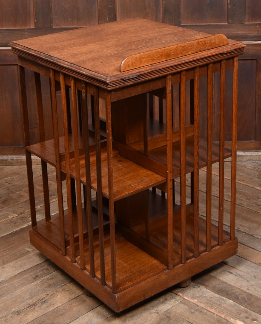 A two-tier freestanding timber revolving bookcase with slatted sides and an angled reading lectern top holding an open volume. View VII