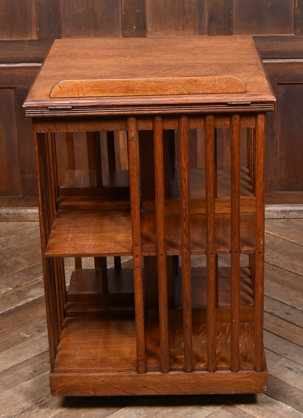 A two-tier freestanding timber revolving bookcase with slatted sides and an angled reading lectern top holding an open volume. View VIII