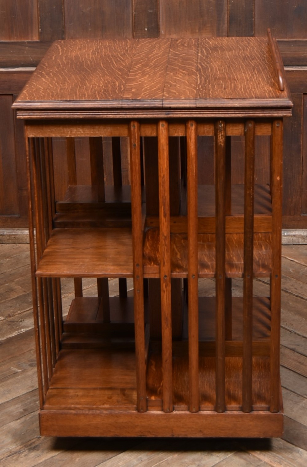 A two-tier freestanding timber revolving bookcase with slatted sides and an angled reading lectern top holding an open volume. View IX