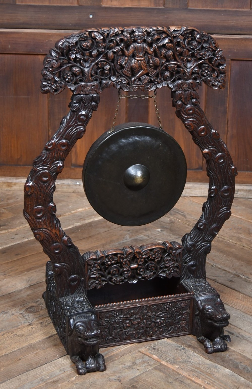An intricately carved dark timber frame with foliage and figure details suspending a dark metal circular nipple gong.