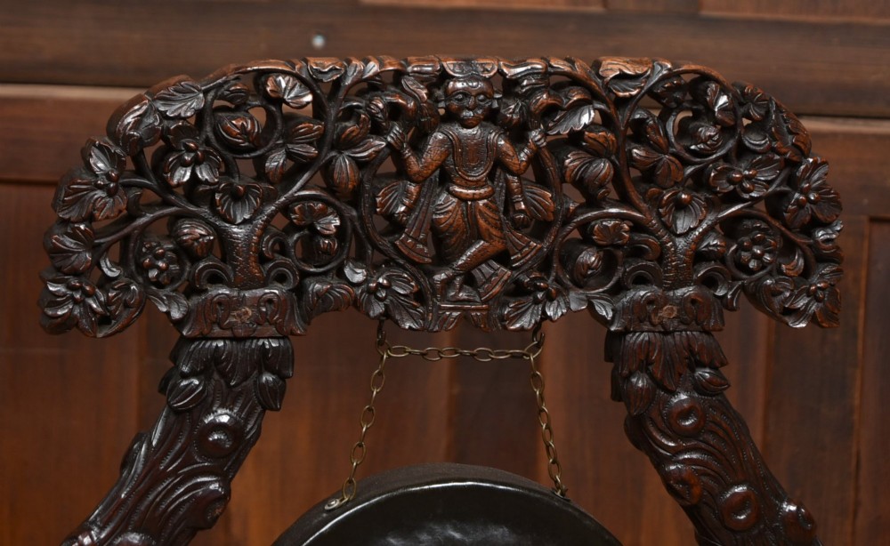 An intricately carved dark timber frame with foliage and figure details suspending a dark metal circular nipple gong. View II