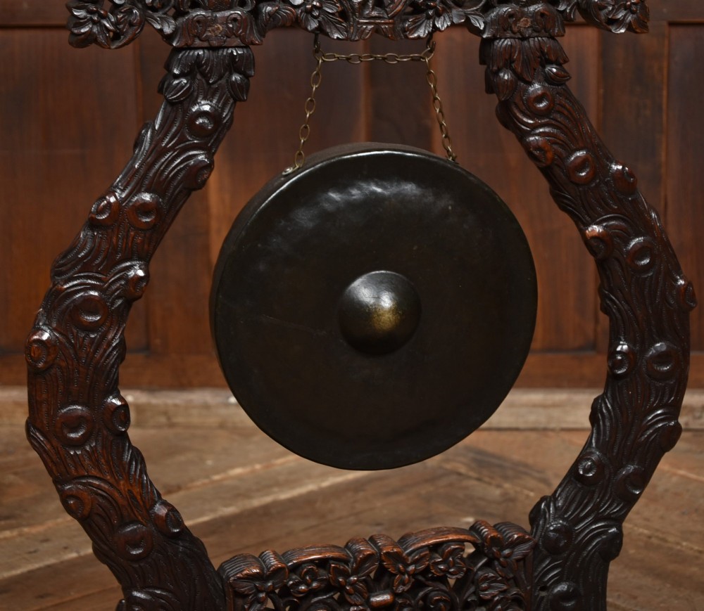 An intricately carved dark timber frame with foliage and figure details suspending a dark metal circular nipple gong. View III