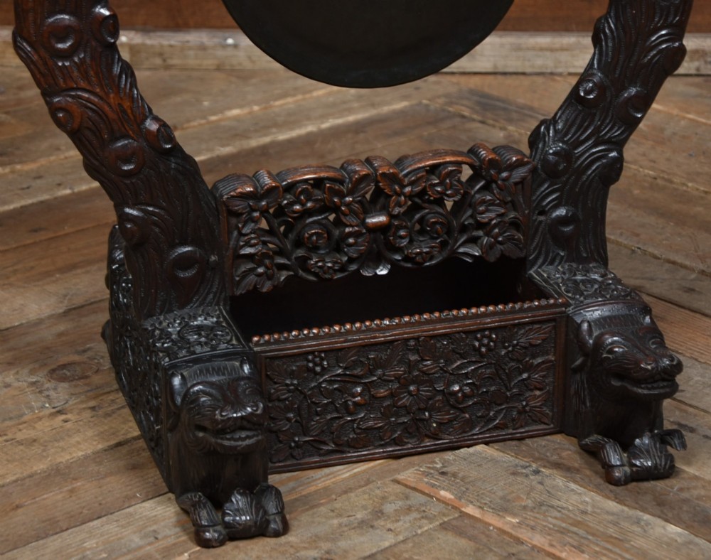 An intricately carved dark timber frame with foliage and figure details suspending a dark metal circular nipple gong. View IV