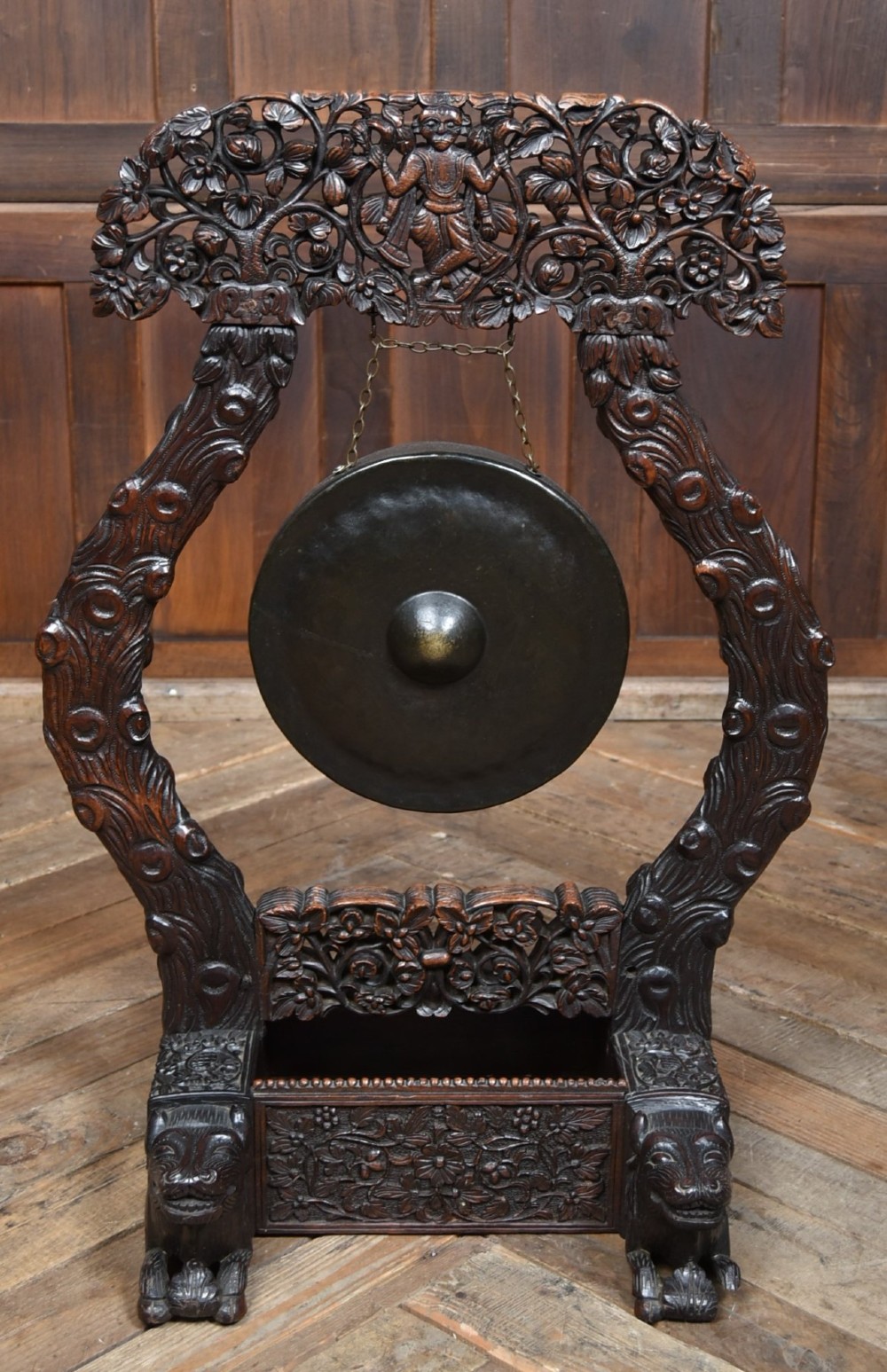 An intricately carved dark timber frame with foliage and figure details suspending a dark metal circular nipple gong. View V