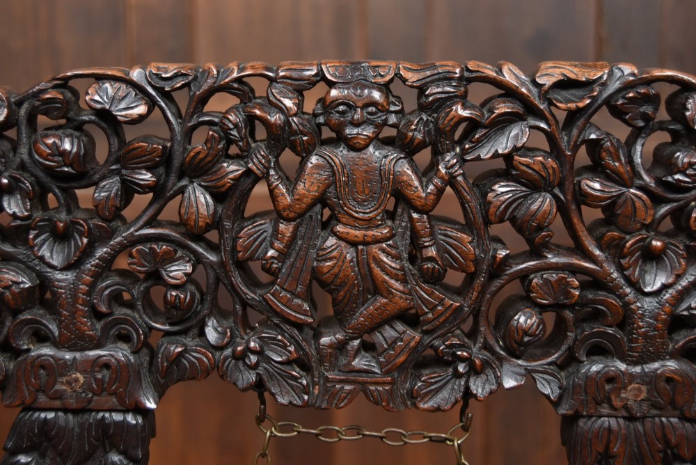 An intricately carved dark timber frame with foliage and figure details suspending a dark metal circular nipple gong. View VI