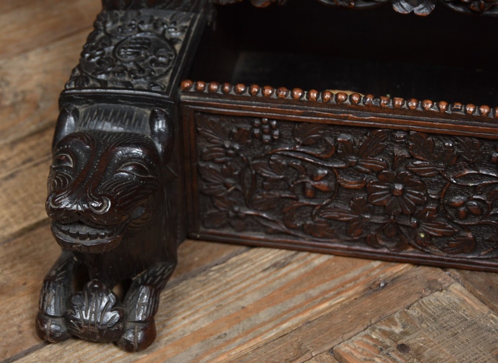 An intricately carved dark timber frame with foliage and figure details suspending a dark metal circular nipple gong. View VII