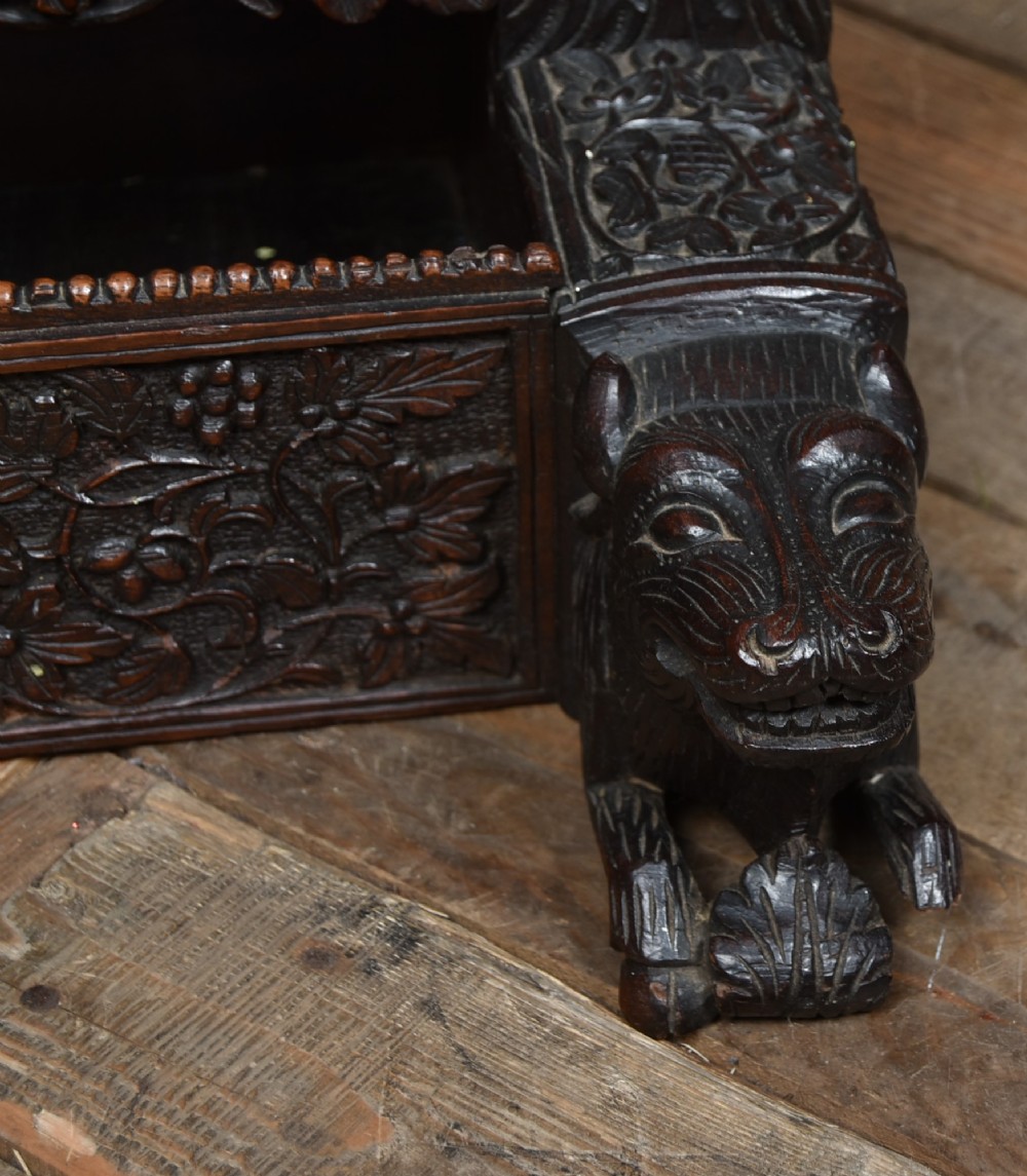 An intricately carved dark timber frame with foliage and figure details suspending a dark metal circular nipple gong. View VIII