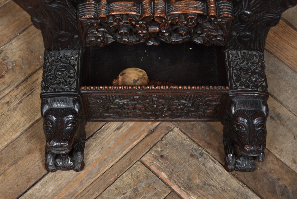 An intricately carved dark timber frame with foliage and figure details suspending a dark metal circular nipple gong. View IX