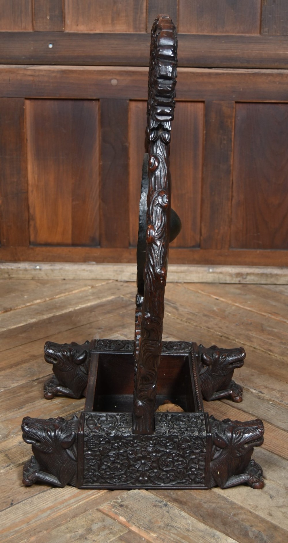 An intricately carved dark timber frame with foliage and figure details suspending a dark metal circular nipple gong. View X