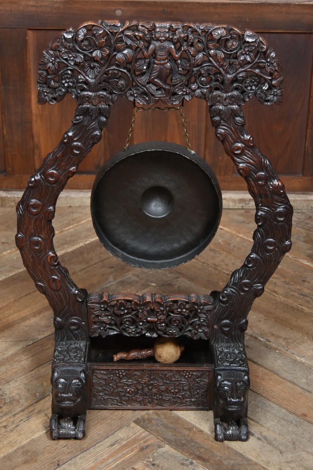 An intricately carved dark timber frame with foliage and figure details suspending a dark metal circular nipple gong. View XI