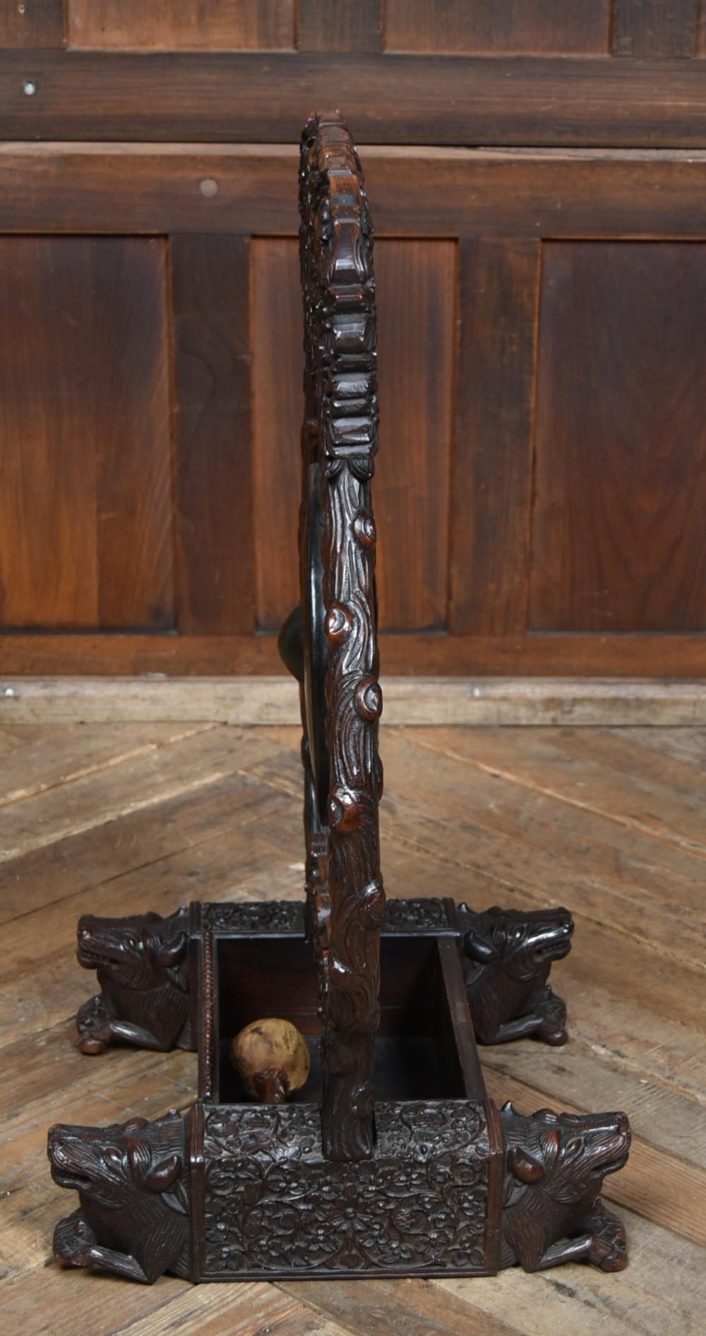 An intricately carved dark timber frame with foliage and figure details suspending a dark metal circular nipple gong. View XII