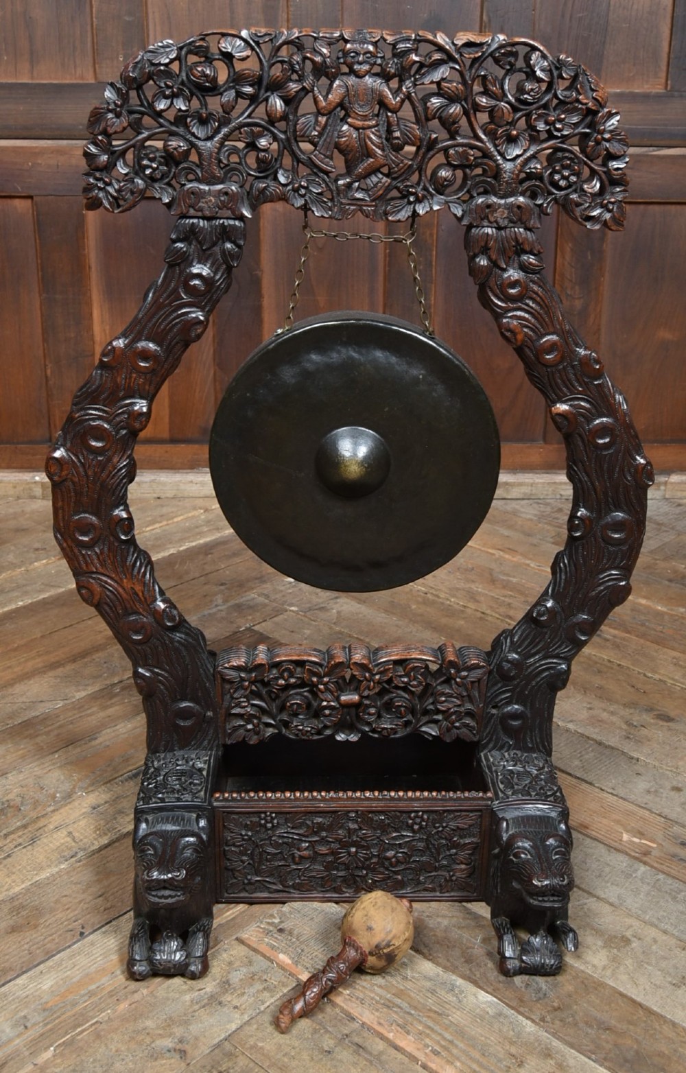 An intricately carved dark timber frame with foliage and figure details suspending a dark metal circular nipple gong. View XIII