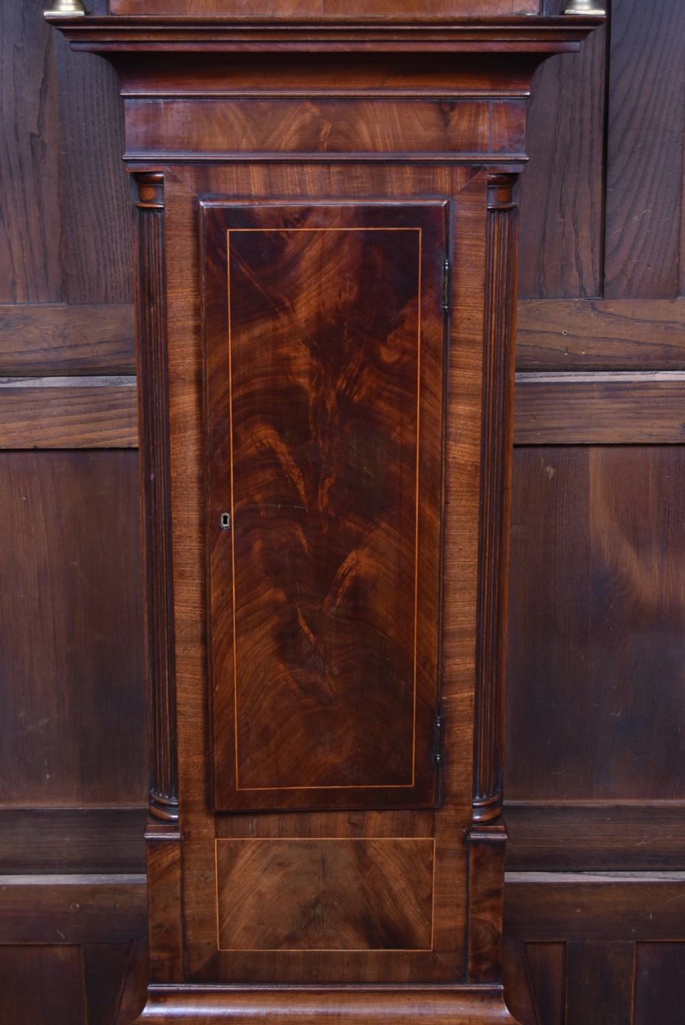 A Scottish mahogany longcase clock featuring a swan-neck pediment with brass finials, a hand-painted dial face, and flame mahogany veneers. View IV
