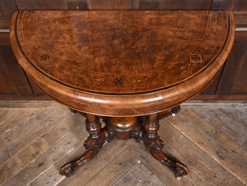 A Victorian burr walnut fold-over games table featuring a semi-circular top, dual turned column pedestal base, carved scroll legs, and ceramic castors. View III