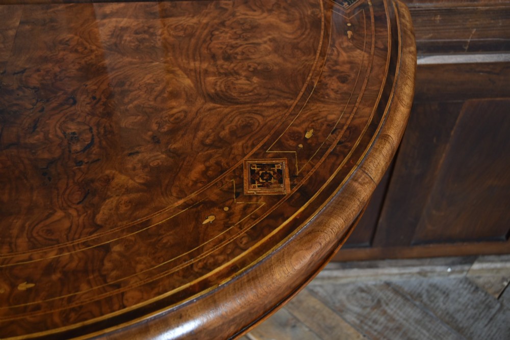 A Victorian burr walnut fold-over games table featuring a semi-circular top, dual turned column pedestal base, carved scroll legs, and ceramic castors. View IV