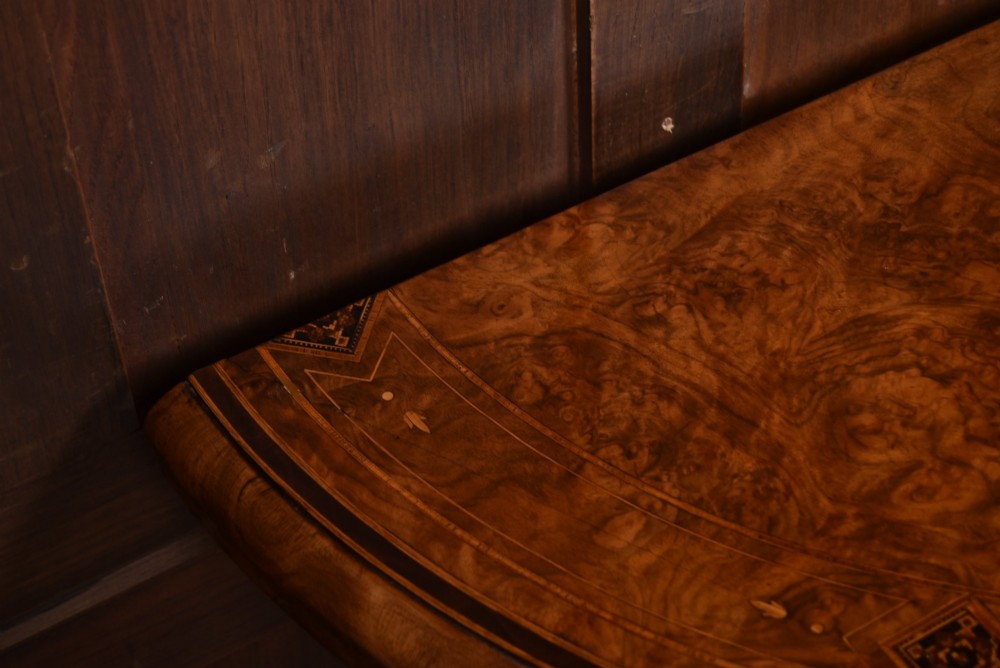 A Victorian burr walnut fold-over games table featuring a semi-circular top, dual turned column pedestal base, carved scroll legs, and ceramic castors. View VI