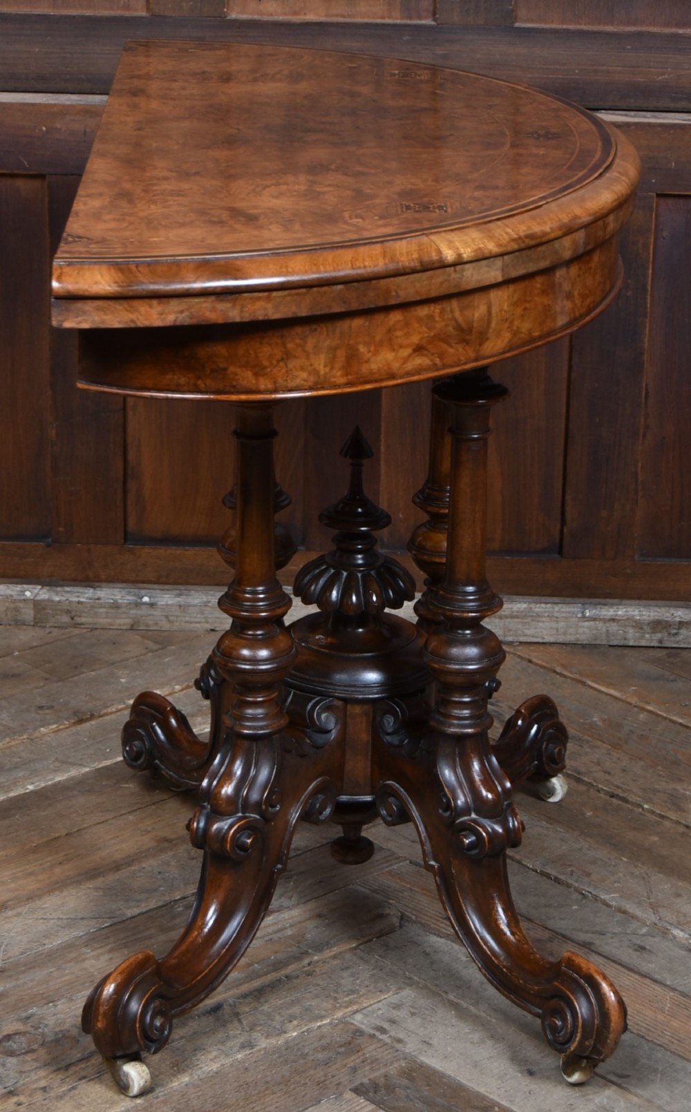 A Victorian burr walnut fold-over games table featuring a semi-circular top, dual turned column pedestal base, carved scroll legs, and ceramic castors. View X
