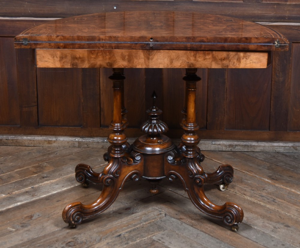 A Victorian burr walnut fold-over games table featuring a semi-circular top, dual turned column pedestal base, carved scroll legs, and ceramic castors. View XI