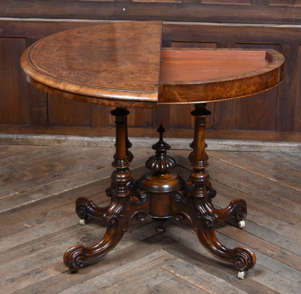 A Victorian burr walnut fold-over games table featuring a semi-circular top, dual turned column pedestal base, carved scroll legs, and ceramic castors. View XIII