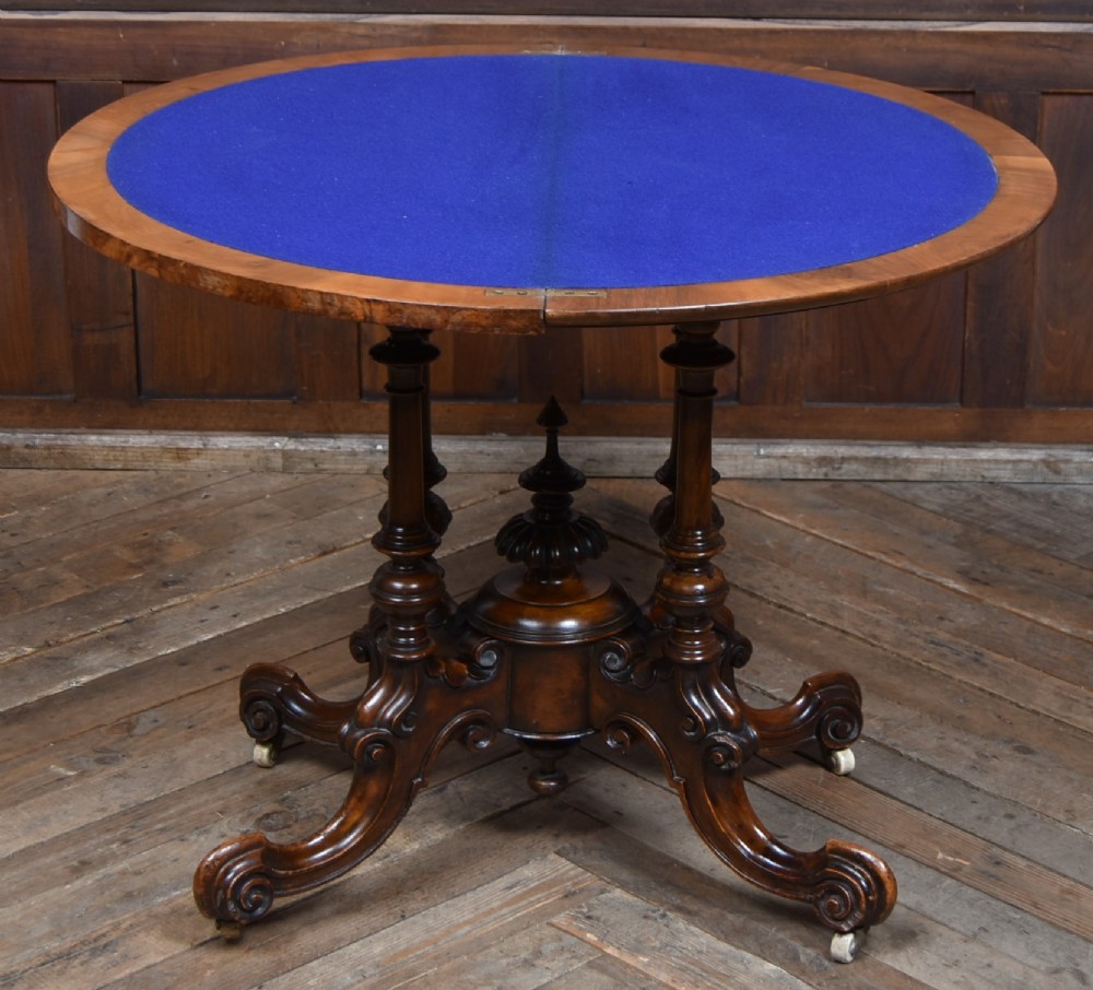 A Victorian burr walnut fold-over games table featuring a semi-circular top, dual turned column pedestal base, carved scroll legs, and ceramic castors. View XIV