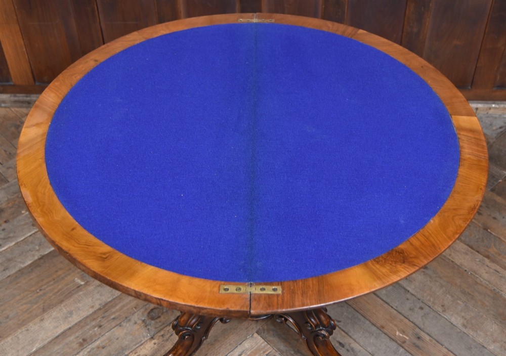 A Victorian burr walnut fold-over games table featuring a semi-circular top, dual turned column pedestal base, carved scroll legs, and ceramic castors. View XV