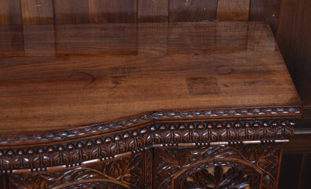A nineteenth-century padouk wood sideboard featuring high-relief foliate hand-carving across four panelled cupboard doors and a breakfront profile. View IV