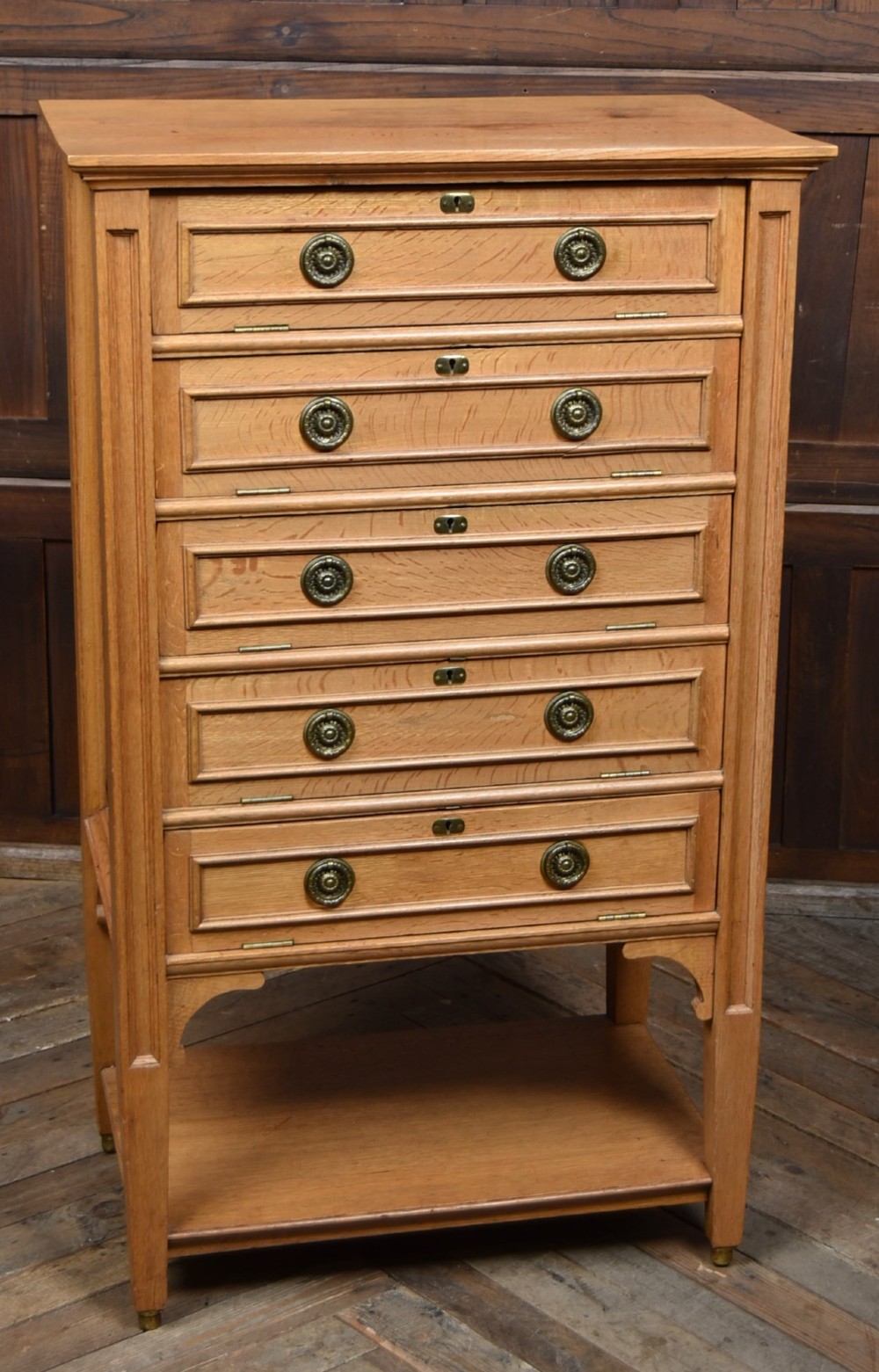 An Edwardian light oak filing cabinet with five stacked fall-front drawers featuring brass ring pulls, supported on square legs with a lower open shelf.