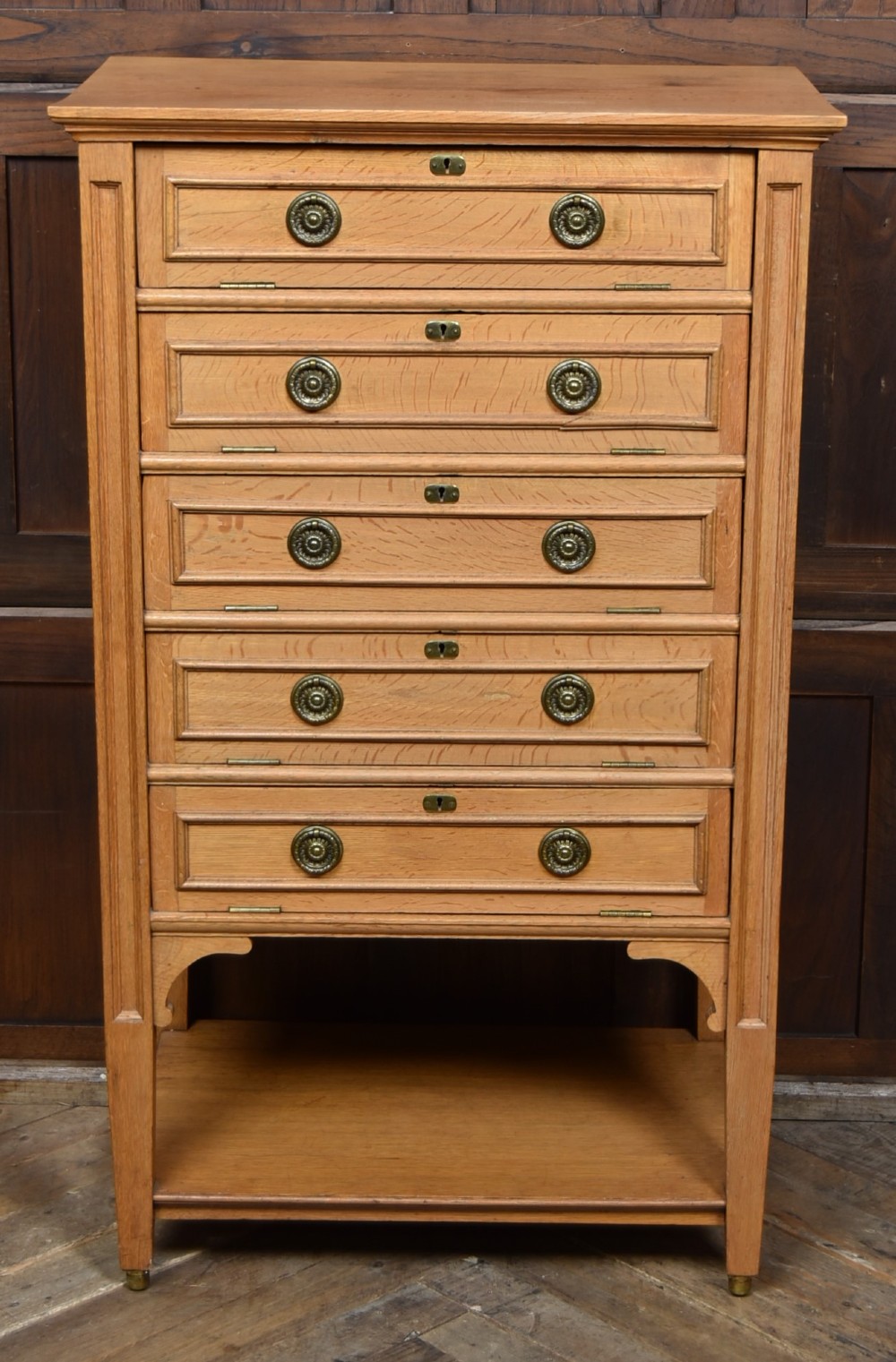 An Edwardian light oak filing cabinet with five stacked fall-front drawers featuring brass ring pulls, supported on square legs with a lower open shelf. View II