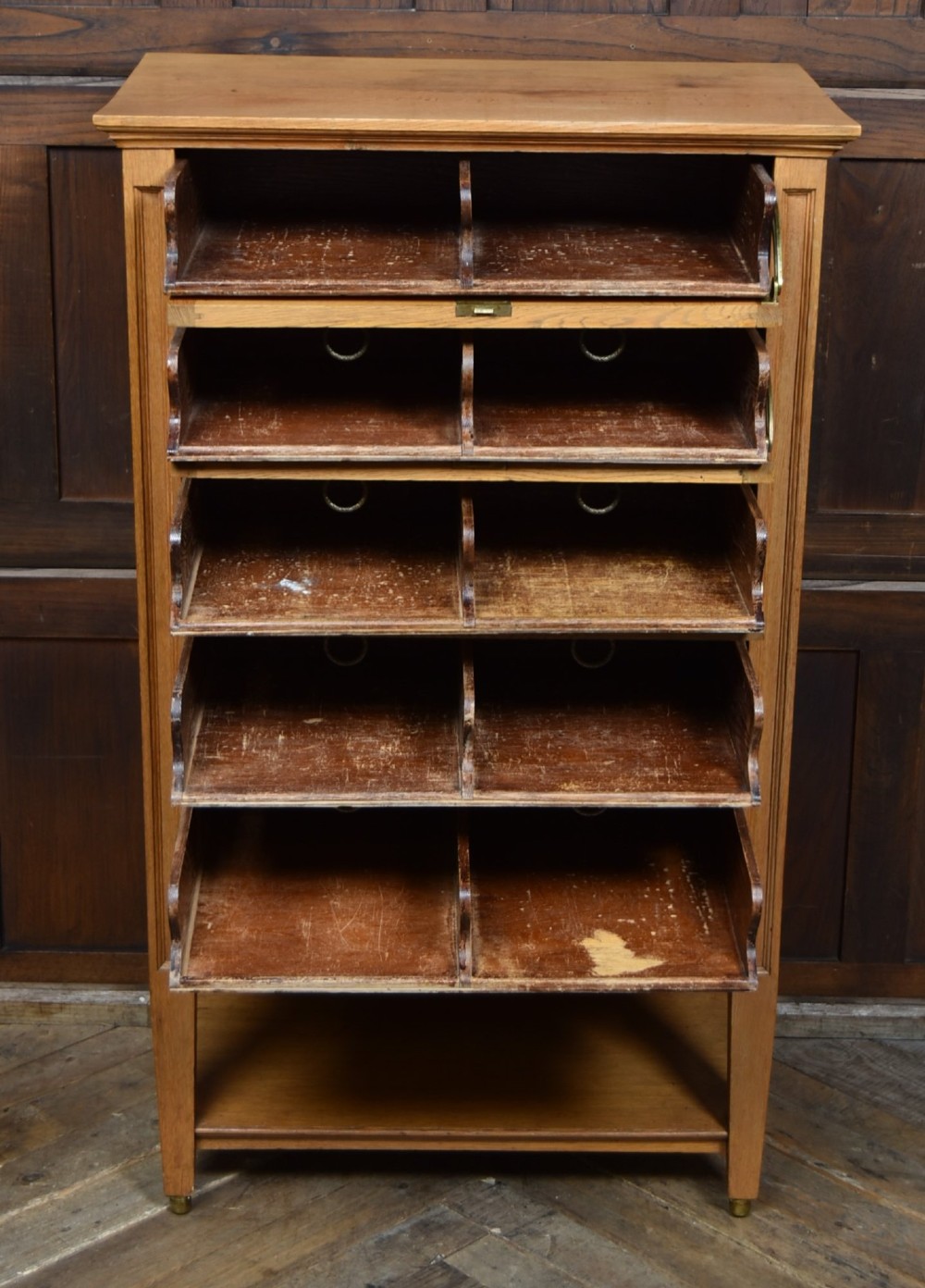 An Edwardian light oak filing cabinet with five stacked fall-front drawers featuring brass ring pulls, supported on square legs with a lower open shelf. View VI