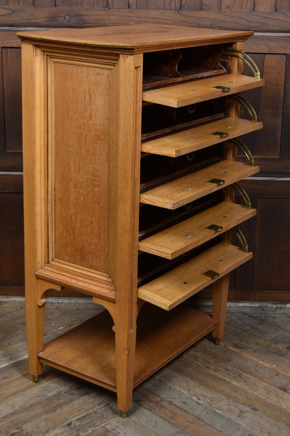 An Edwardian light oak filing cabinet with five stacked fall-front drawers featuring brass ring pulls, supported on square legs with a lower open shelf. View VIII