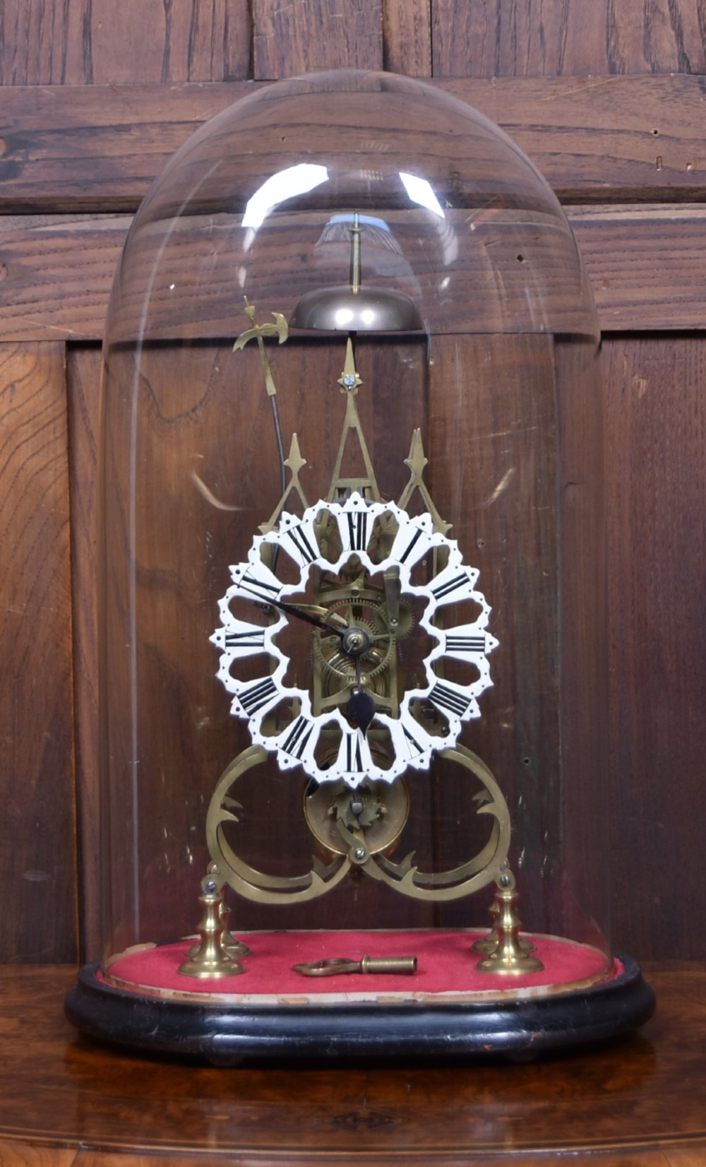 A Victorian brass skeleton clock with an openwork pierced dial and top bell, enclosed within a glass dome on an ebonised wooden base.