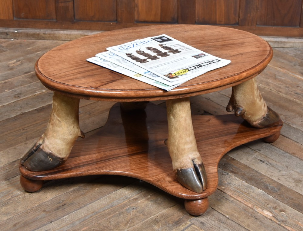 An oval polished wooden coffee table supported by four taxidermy hooves standing on a shaped wooden base with ball feet.