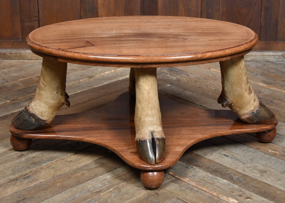 An oval polished wooden coffee table supported by four taxidermy hooves standing on a shaped wooden base with ball feet. View VI
