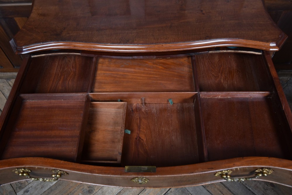 A Georgian mahogany chest of drawers featuring a serpentine front, four graduated drawers with ornate brass handles, and carved canted corners. View V