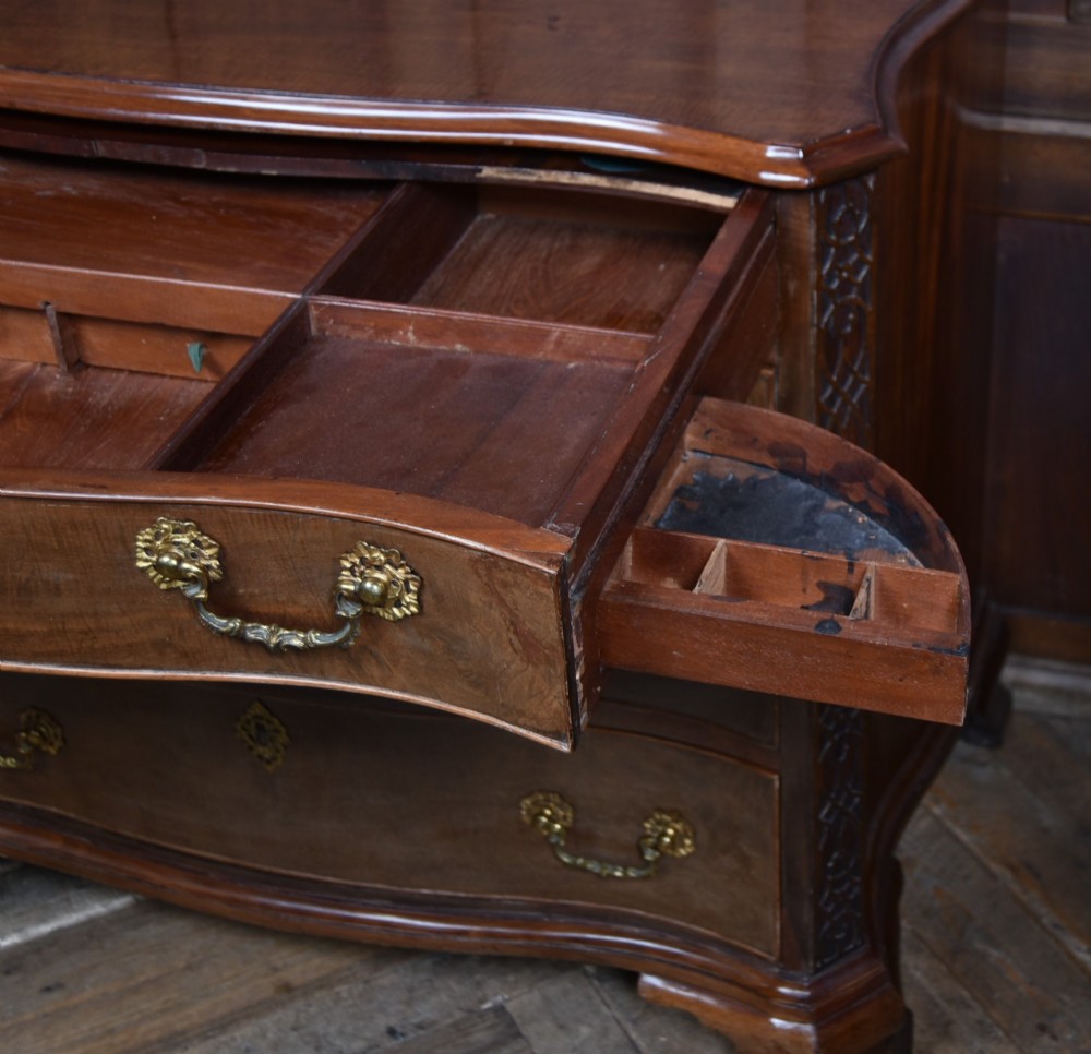 A Georgian mahogany chest of drawers featuring a serpentine front, four graduated drawers with ornate brass handles, and carved canted corners. View VI