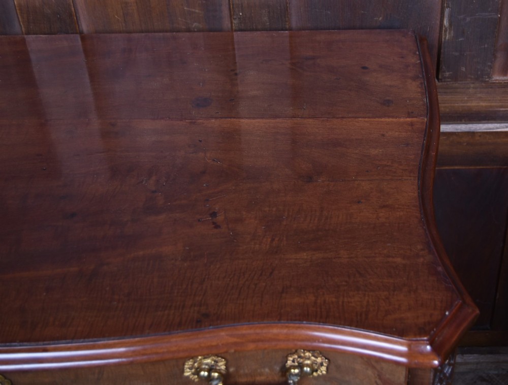 A Georgian mahogany chest of drawers featuring a serpentine front, four graduated drawers with ornate brass handles, and carved canted corners. View VII