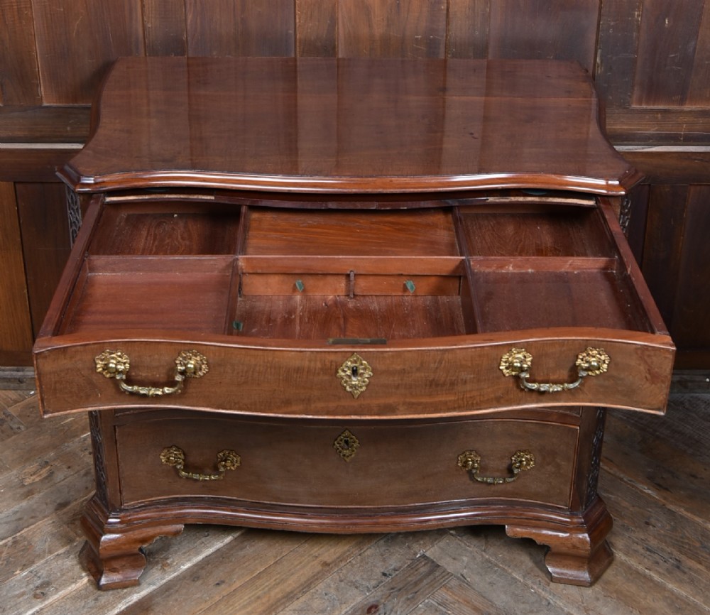 A Georgian mahogany chest of drawers featuring a serpentine front, four graduated drawers with ornate brass handles, and carved canted corners. View XIII