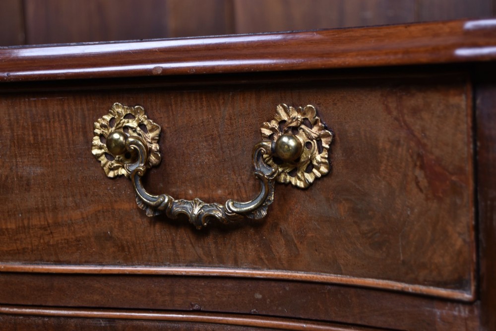 A Georgian mahogany chest of drawers featuring a serpentine front, four graduated drawers with ornate brass handles, and carved canted corners. View XIV