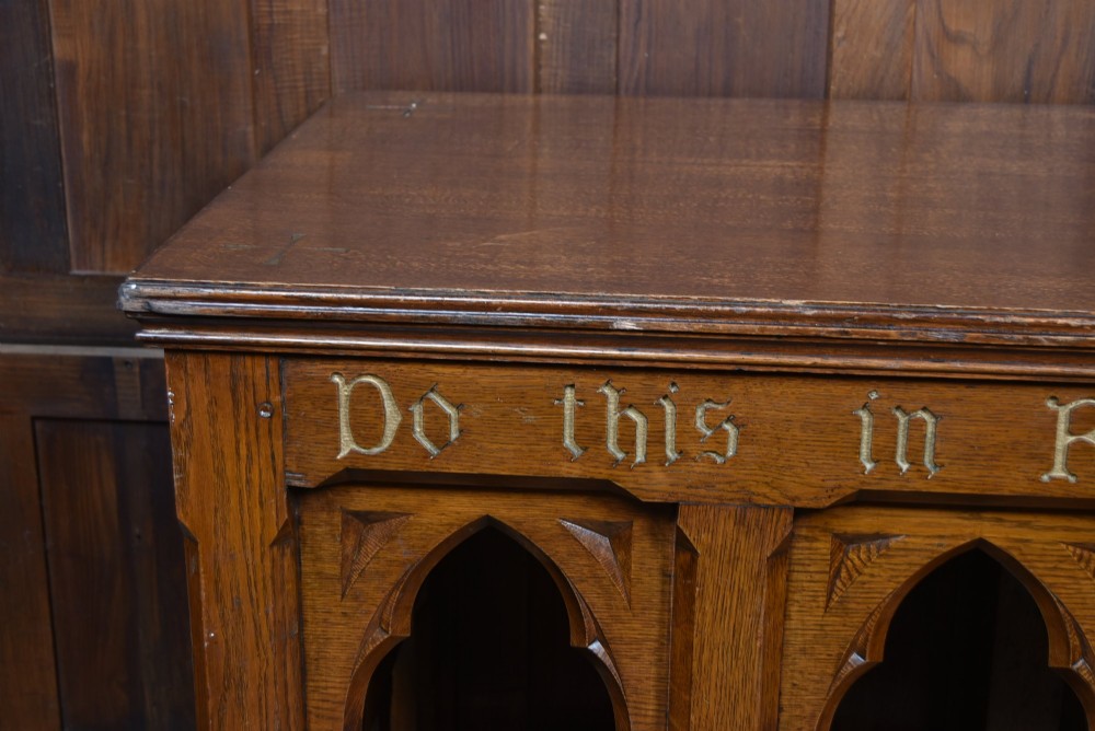 A Victorian oak church communion table with carved Gothic arch panels and a carved inscription reading Do this in Remembrance of me along the top frieze. View II