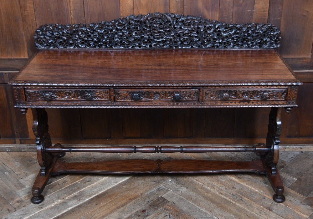 A dark brown padouk wood side table featuring a deeply carved floral gallery back, three carved drawer fronts, and trestle legs connected by a turned stretcher.