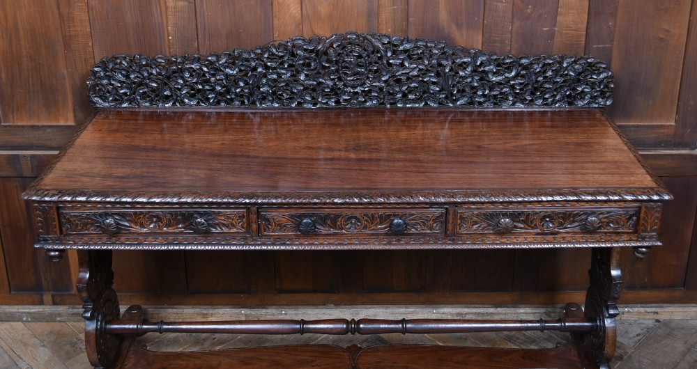 A dark brown padouk wood side table featuring a deeply carved floral gallery back, three carved drawer fronts, and trestle legs connected by a turned stretcher. View II