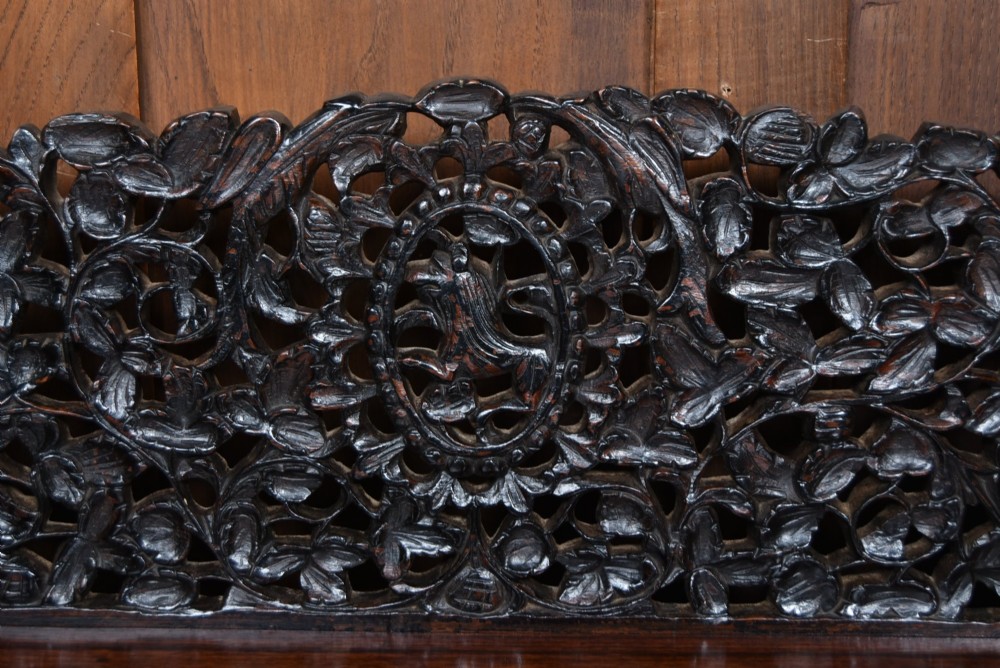 A dark brown padouk wood side table featuring a deeply carved floral gallery back, three carved drawer fronts, and trestle legs connected by a turned stretcher. View III