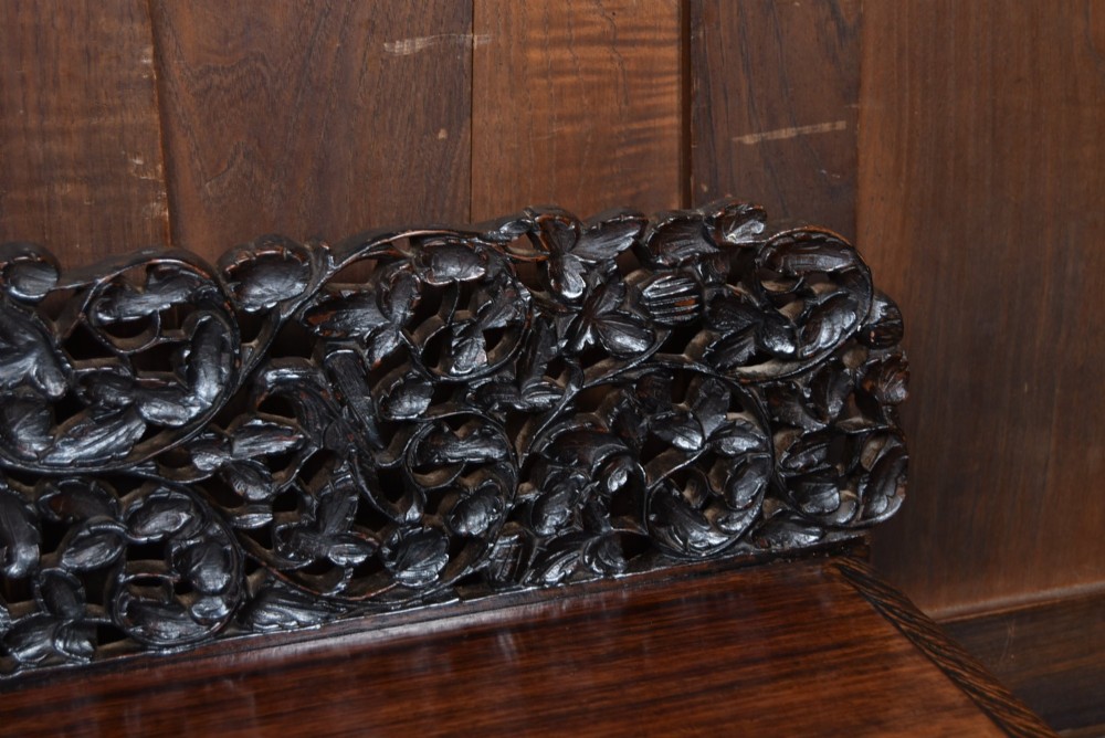 A dark brown padouk wood side table featuring a deeply carved floral gallery back, three carved drawer fronts, and trestle legs connected by a turned stretcher. View IV
