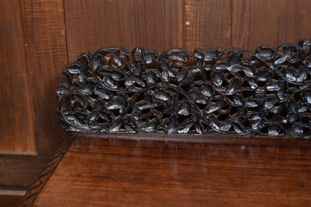 A dark brown padouk wood side table featuring a deeply carved floral gallery back, three carved drawer fronts, and trestle legs connected by a turned stretcher. View V