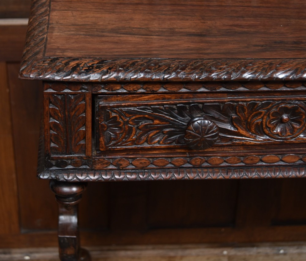 A dark brown padouk wood side table featuring a deeply carved floral gallery back, three carved drawer fronts, and trestle legs connected by a turned stretcher. View VI