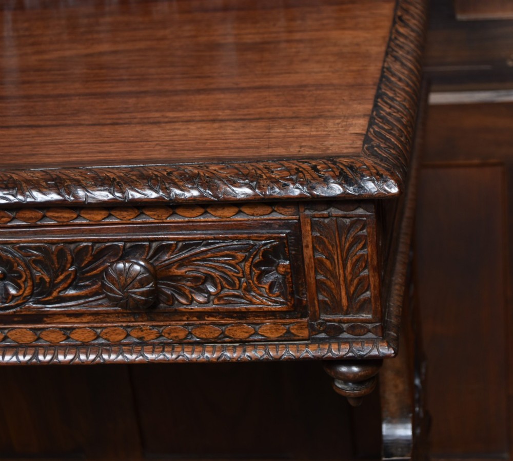 A dark brown padouk wood side table featuring a deeply carved floral gallery back, three carved drawer fronts, and trestle legs connected by a turned stretcher. View VII