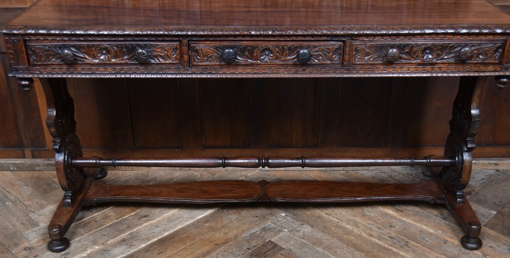 A dark brown padouk wood side table featuring a deeply carved floral gallery back, three carved drawer fronts, and trestle legs connected by a turned stretcher. View VIII