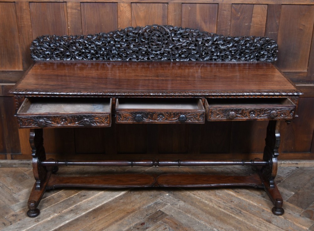 A dark brown padouk wood side table featuring a deeply carved floral gallery back, three carved drawer fronts, and trestle legs connected by a turned stretcher. View IX