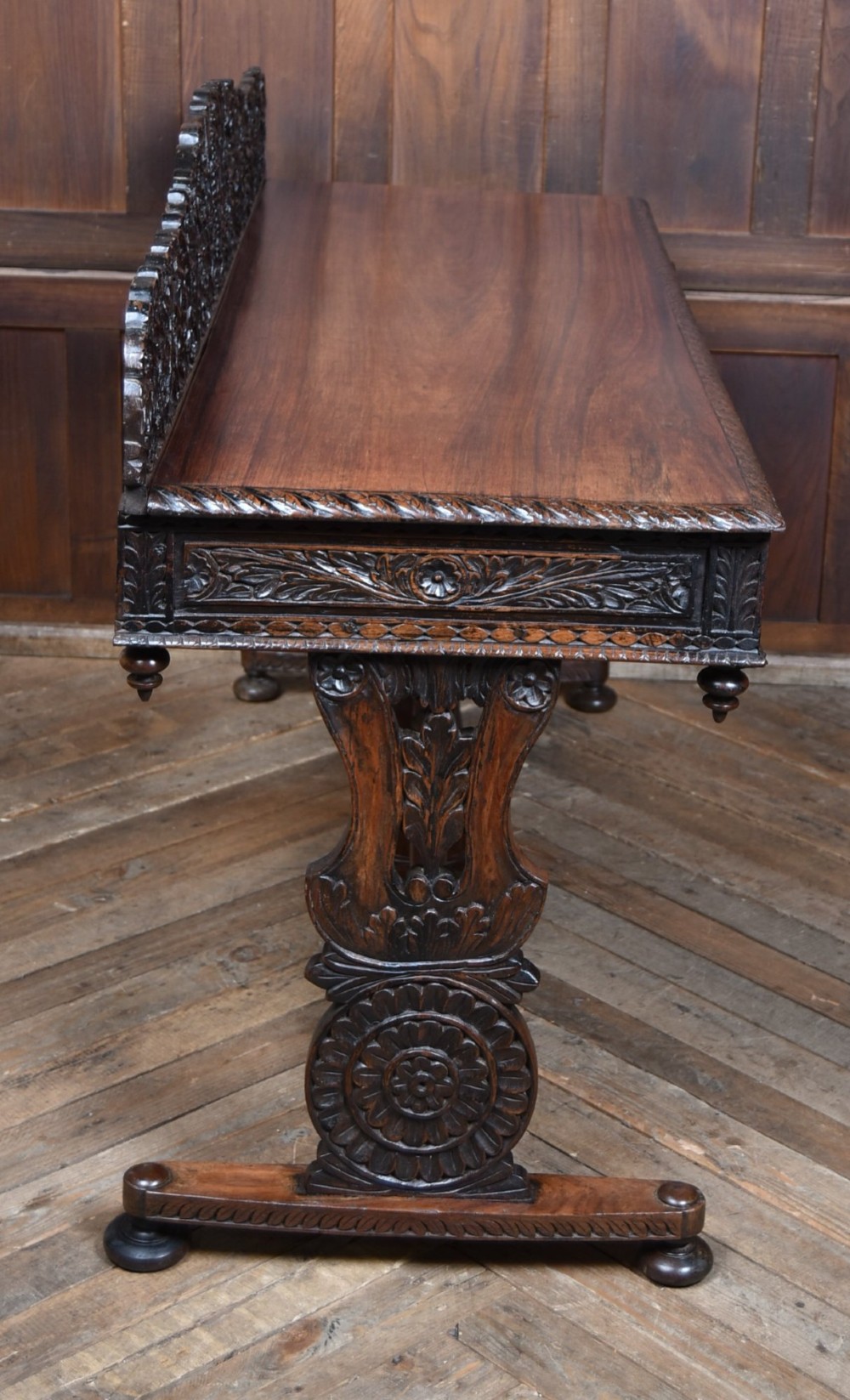 A dark brown padouk wood side table featuring a deeply carved floral gallery back, three carved drawer fronts, and trestle legs connected by a turned stretcher. View X